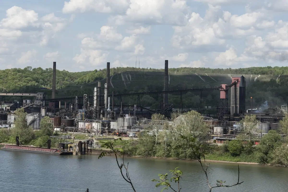 Steel plant smokestacks, conveyors and tanks along a river below wooded hills